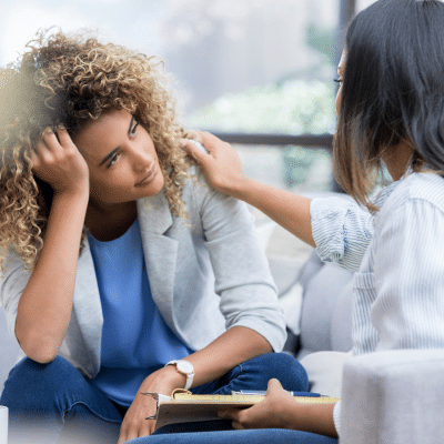 A woman sitting on a couch holding her head while another woman holds her shoulder, representing mind-body connection